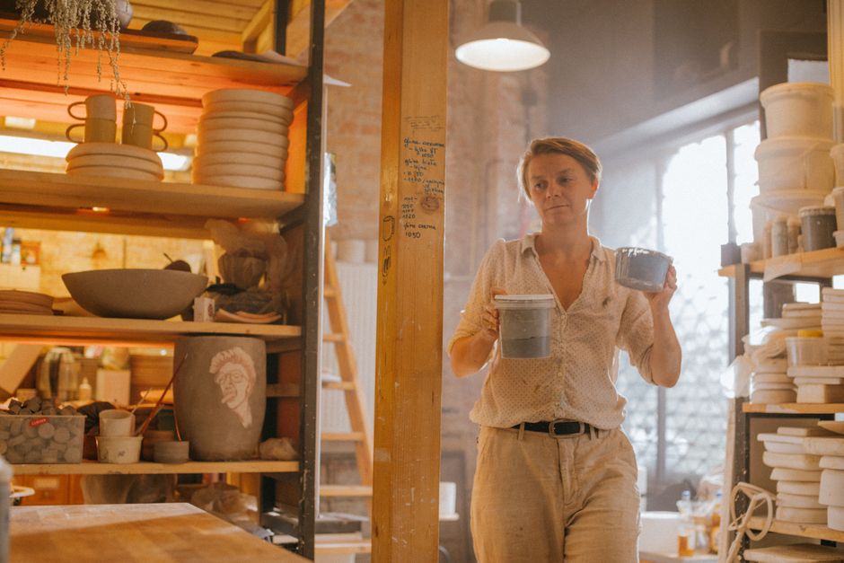 A woman artist holding ceramic bowls in a sunlit workshop, surrounded by shelves filled with pottery.