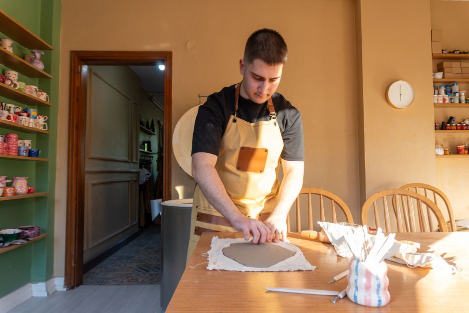 A potter in an apron works with clay in a sunlit studio, surrounded by pottery tools.